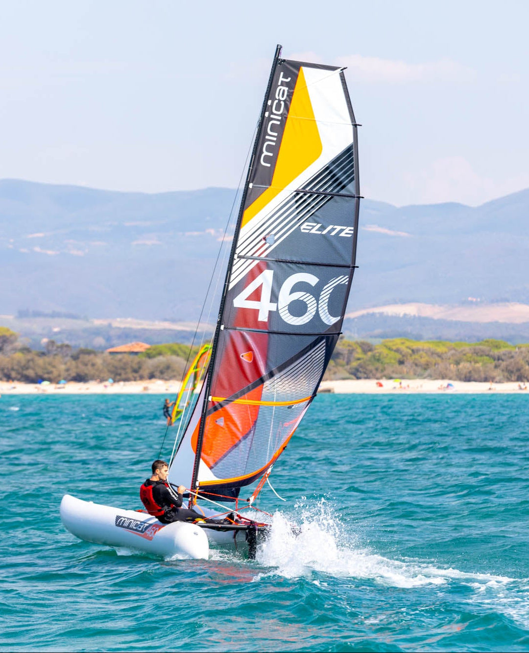 Person sailing MiniCat on a blue ocean with a colorful sail, mountains in the background.