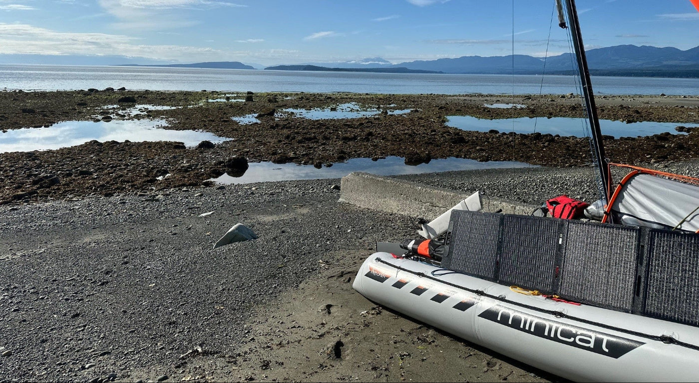 Inflatable catamaran MiniCat on a beach with Remigo fitted and solar charging with a scenic background of water and mountains.