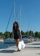 Woman in a black dress holding white bags on a dock with sailboats in the background