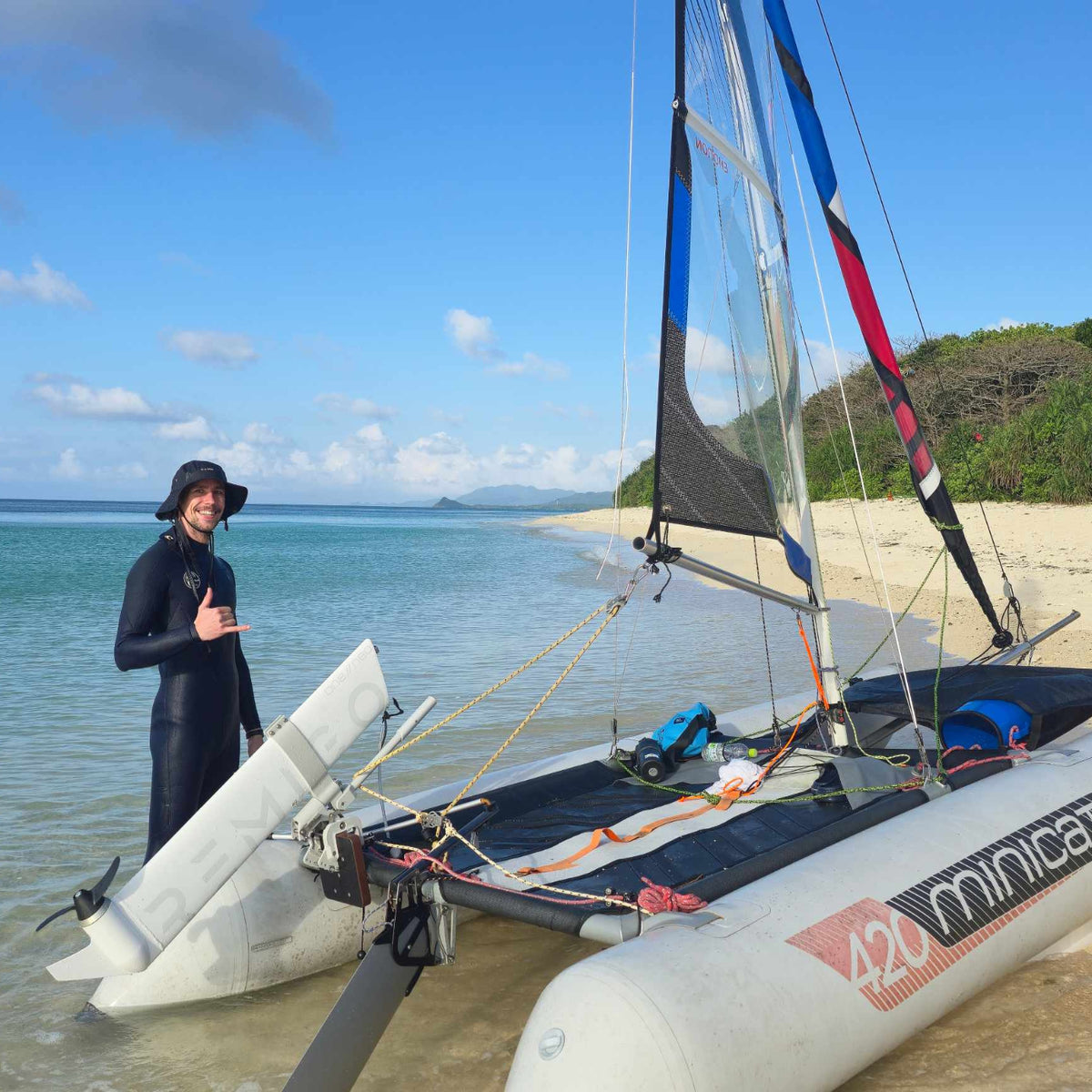 A person in a black wetsuit stands beside a white Minicar catamaran on a sandy beach with blue water and green hills in the background.