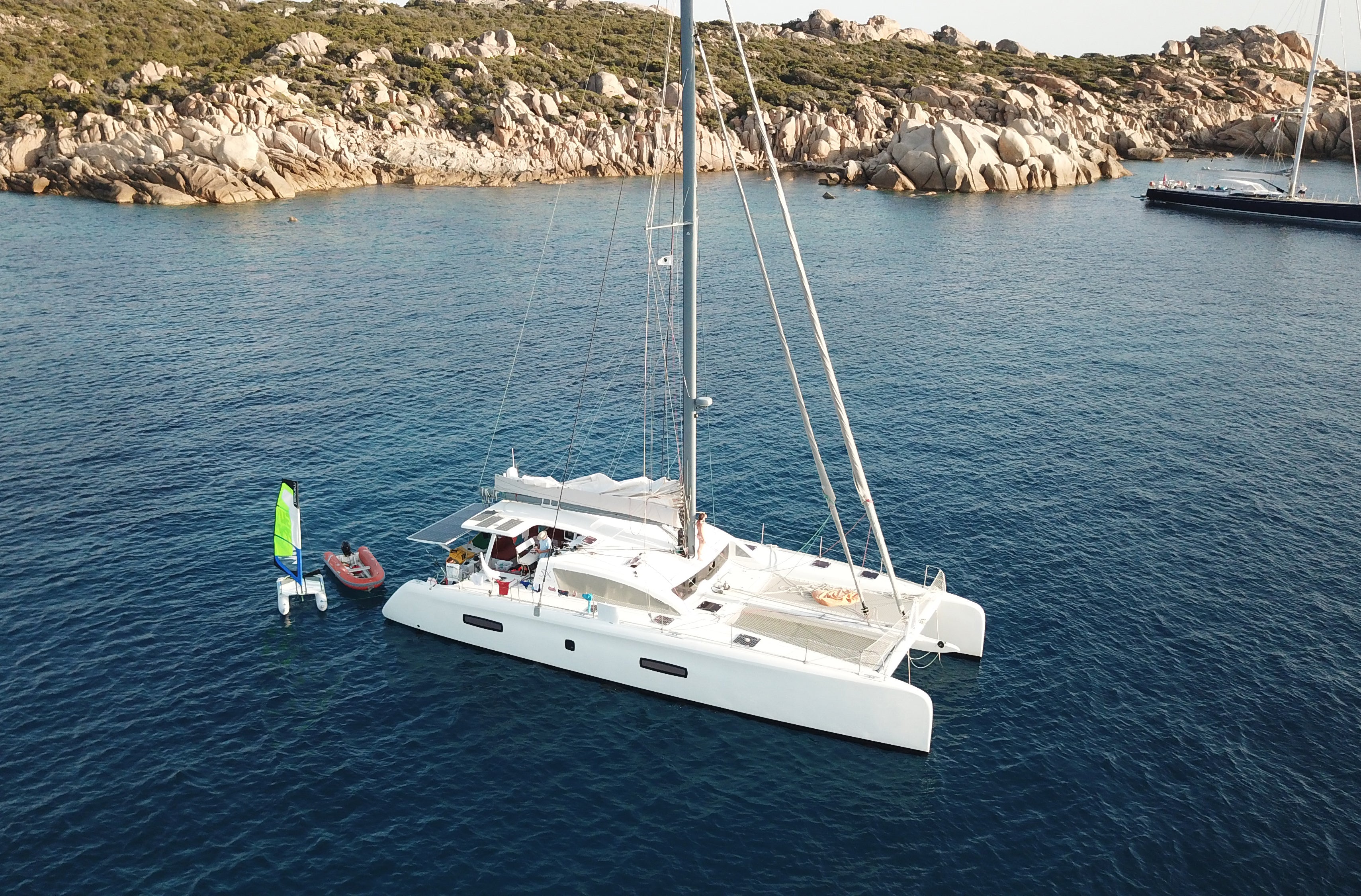 White catamaran sailing on a blue sea with rocky cliffs in the background and MiniCat GUPPY next to it