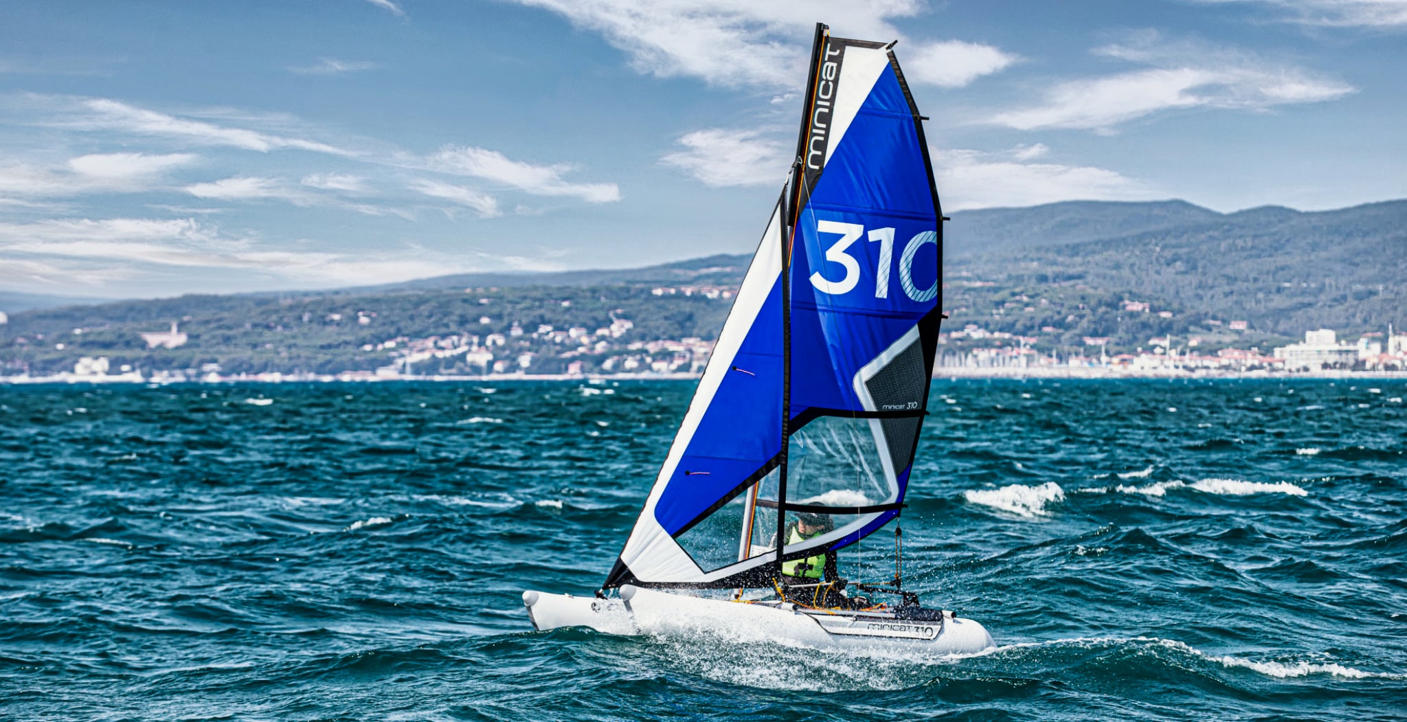 Sailboat with a blue sail featuring the number 310 on a body of water with a cityscape in the background.