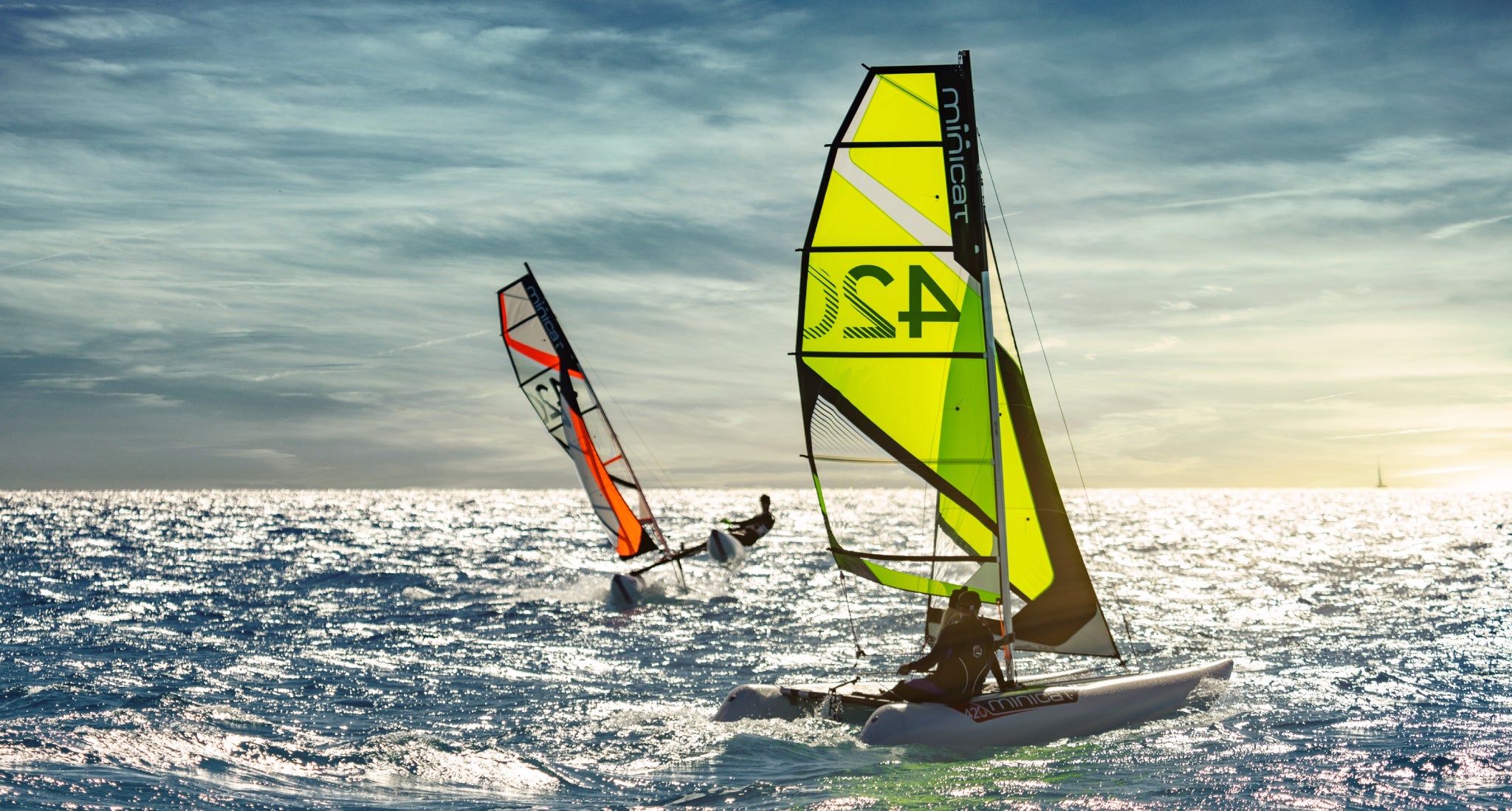 Two sailing catamarans on the water with colorful sails against a cloudy sky.
