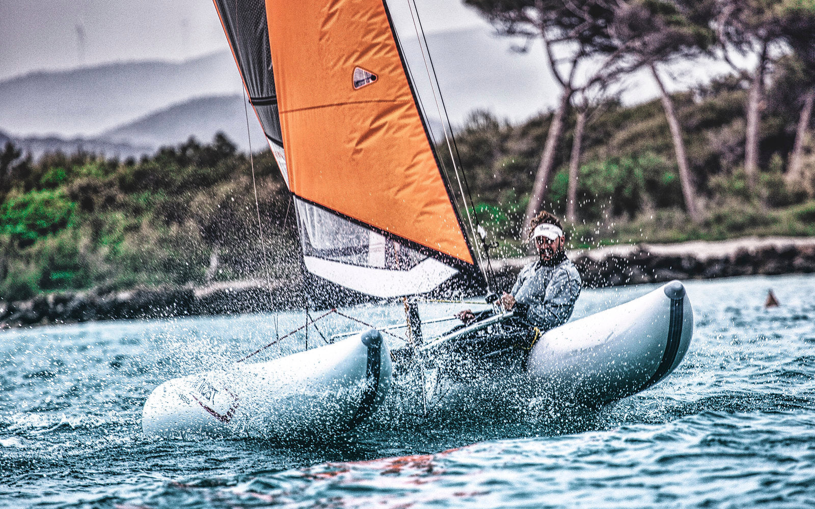 Person sailing on a small inflatable boat MiniCat with an orange sail in a coastal area.