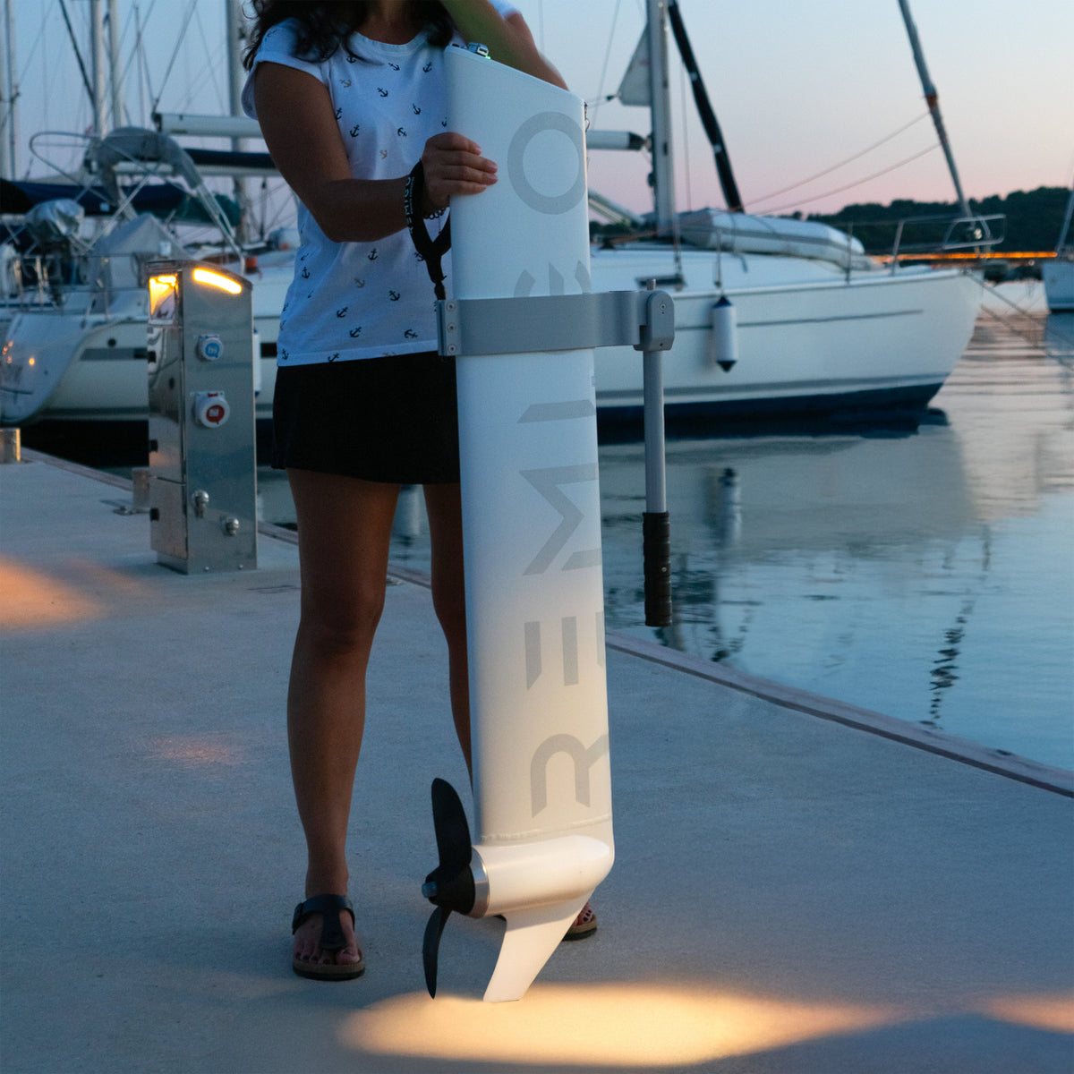 Person holding a white REMIGO electric outboard on a dock with boats in the background.