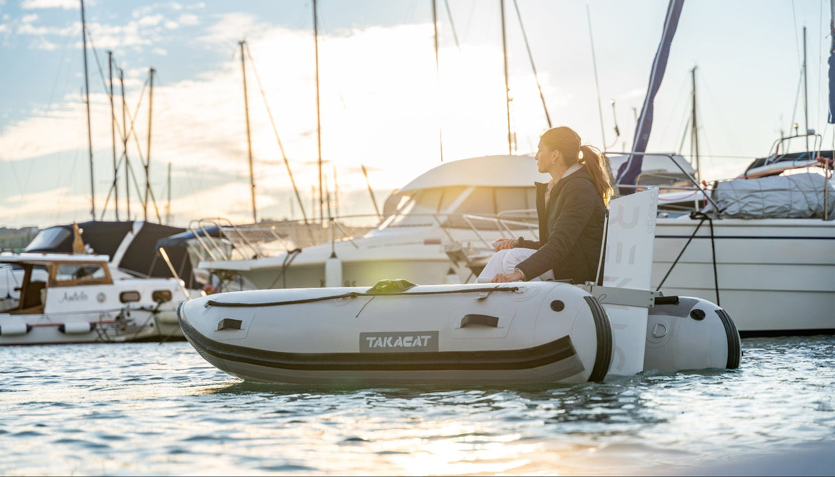 A woman sits in a white and gray Takakat inflatable dinghy on calm water at sunset, with boats and masts in the background.
