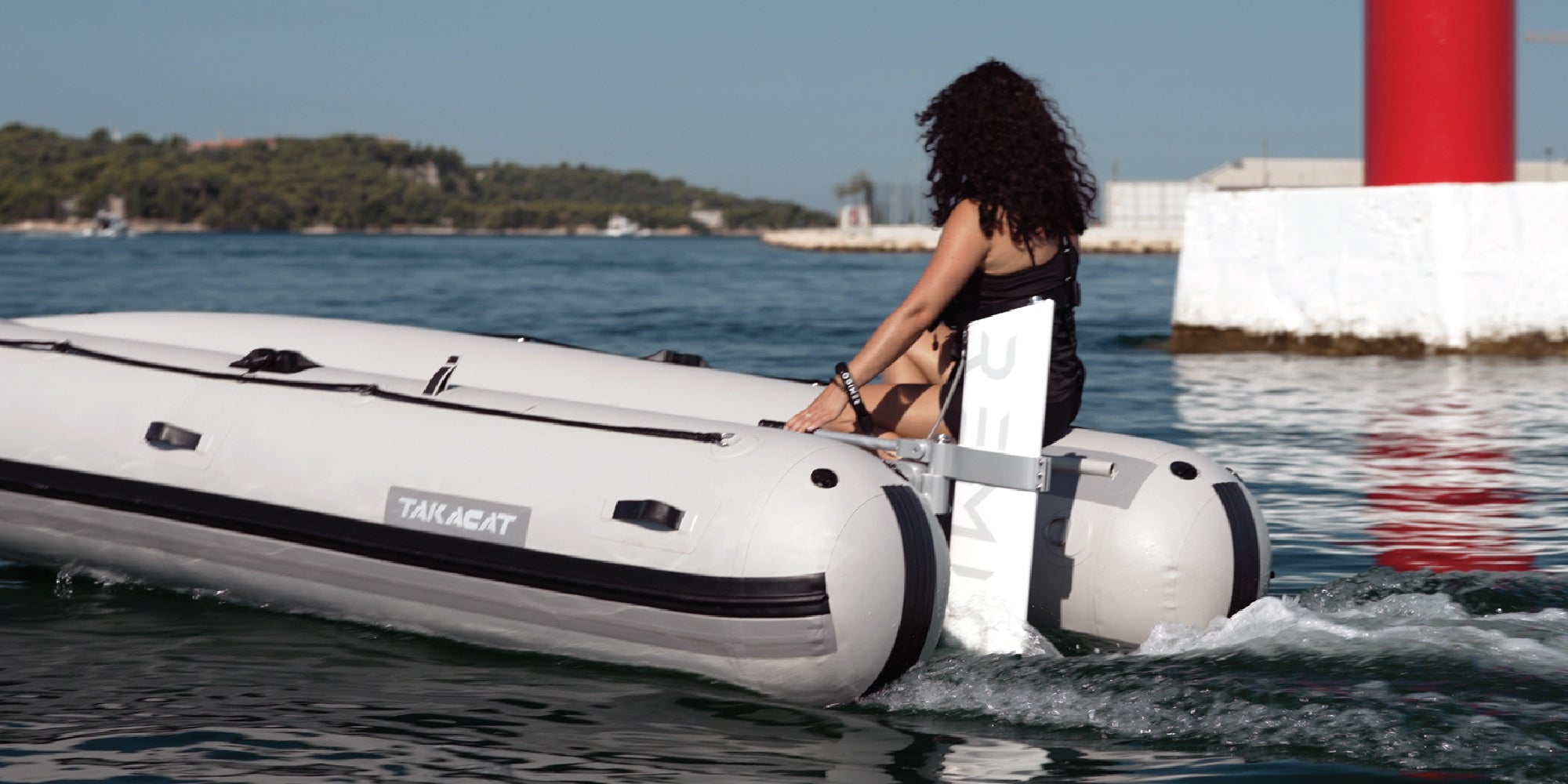 Woman sitting on an inflatable boat with a Remigo electric outboard, surrounded by water and a lighthouse.