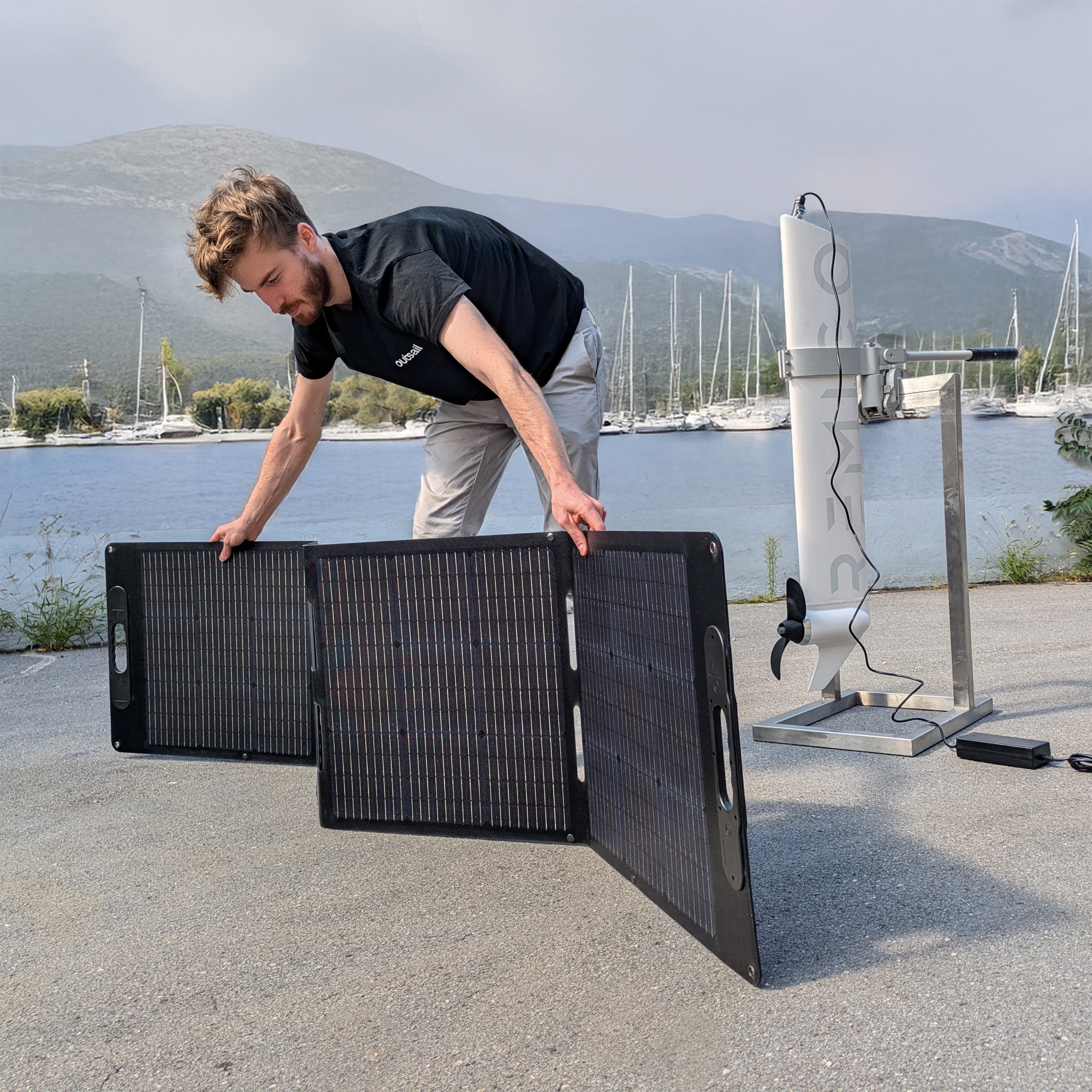 Person setting up a portable solar panel by a waterfront with sailboats.