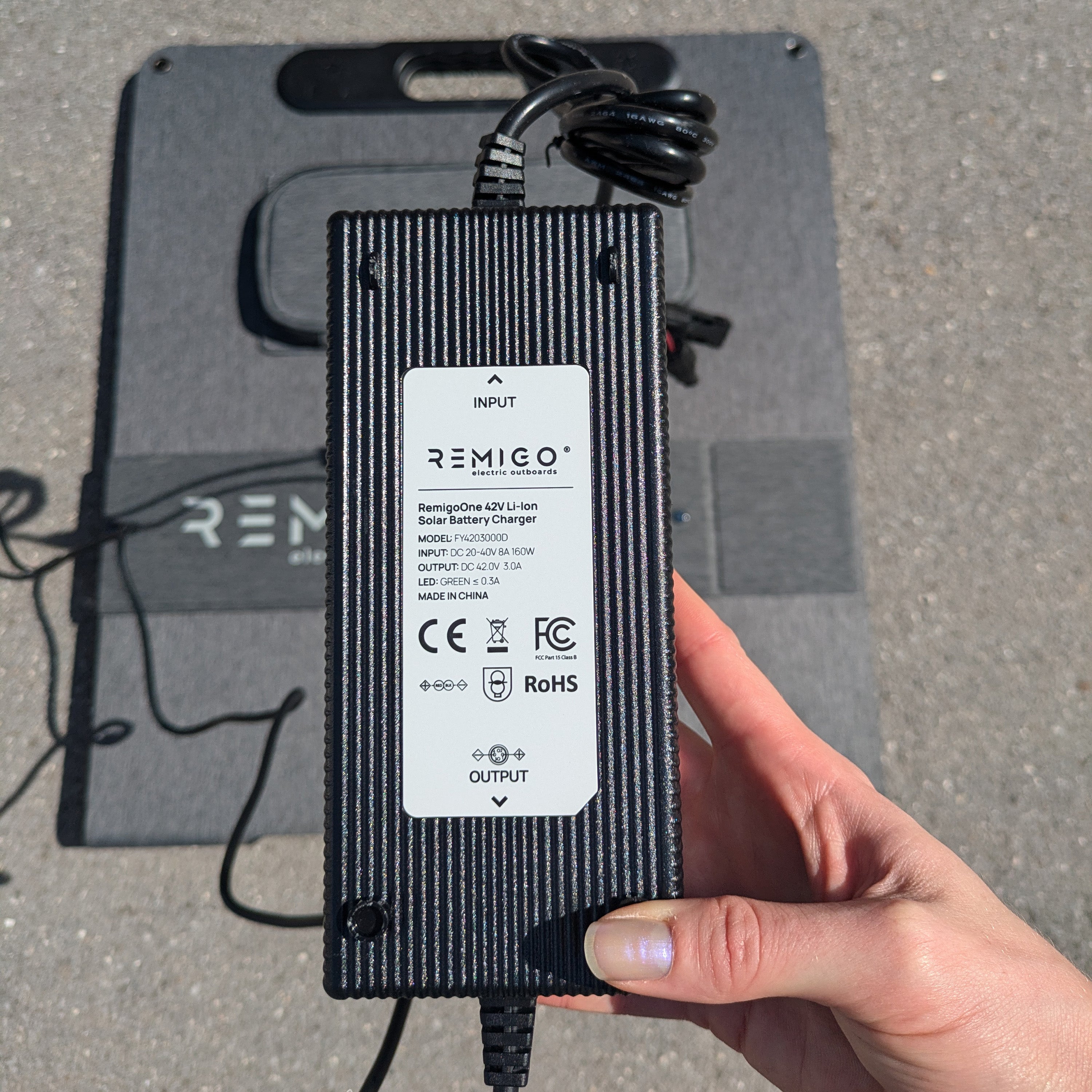 Black power adapter with REMICO branding held by a hand on a gray surface.